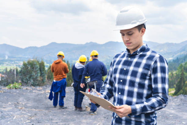Young mining engineering student looking at a spreadsheet held in his hands while young miners and students are in the open pit mine.